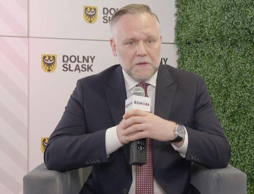 Dawid Jackiewicz podczas wystąpienia publicznego, ubrany w garnitur, gestykulujący, na tle rozmazanej sali konferencyjnej.