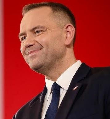 Karol Nawrocki podczas wystąpienia publicznego, na tle symbolizującym jego zaangażowanie w upamiętnianie historii.