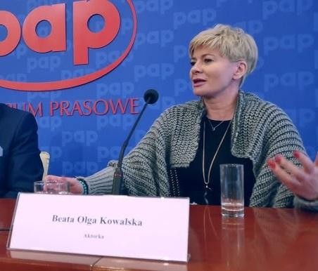 Beata Olga Kowalska na scenie teatralnej, w dynamicznej pozie, wyrażająca silne emocje, z dramatycznym oświetleniem i rozmytym tłem scenografii.