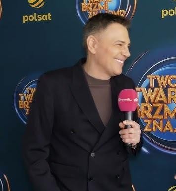 Krzysztof Ibisz prowadzący program telewizyjny, uśmiechnięty i pełen energii, w tle jasne studio z ekranami.